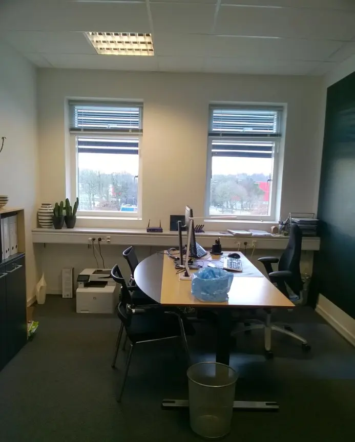 Kontor med to skriveborde, hver med computer, skrivebordslampe og stol samt en papirkurv og to vinduer med gardiner