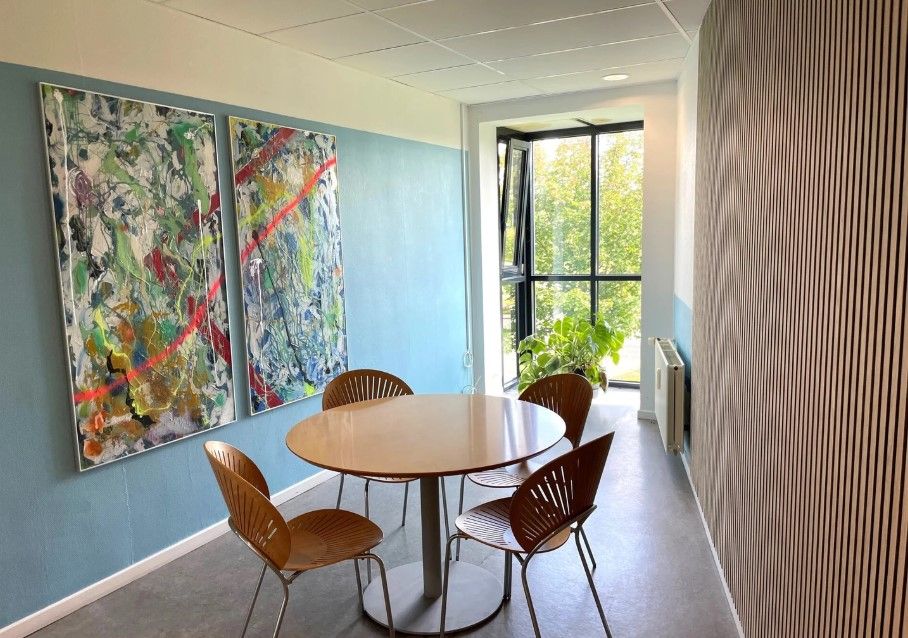 Kontorlokaler med rundt bord og fire stole samt to malerier med abstrakt maleri på blå væg