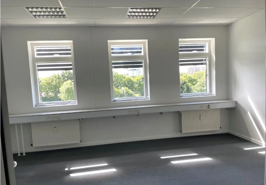 Kontor med tre vinduer, hvide vægge, gulvtæppe og radiatorer under vinduer. Lysstråler trænger ind gennem vinduer og skaber