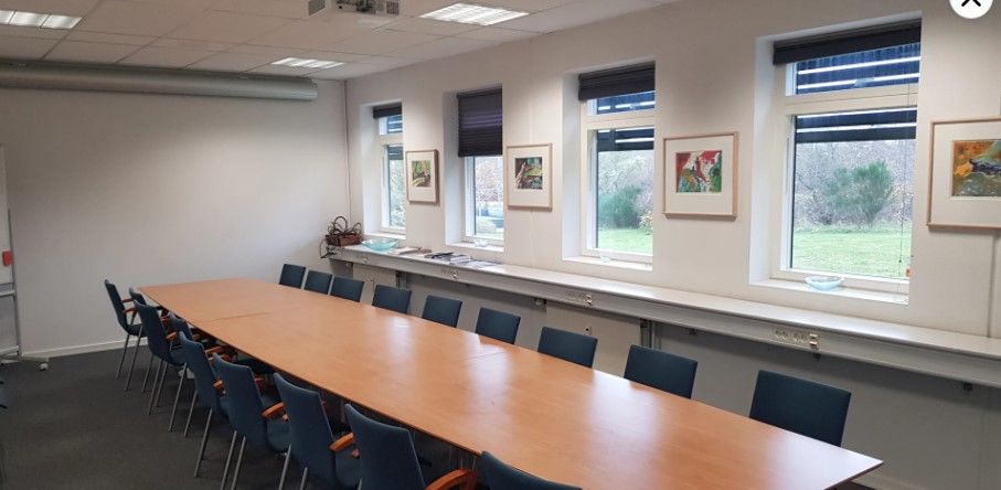 Kontor til leje med bord og stole samt malerier på væggen og vinduer med udsigt til græsarealer.