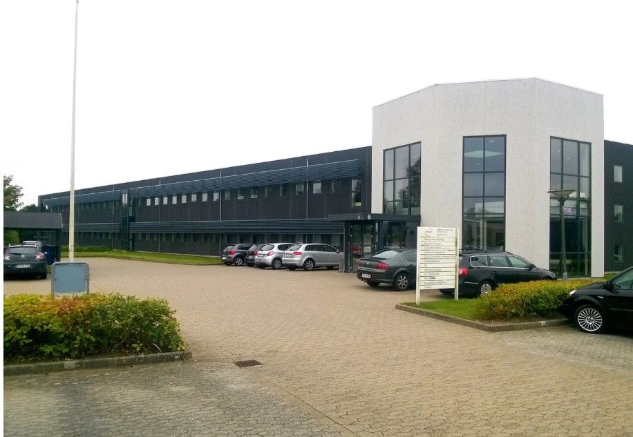 I dette billede er der en moderne bygning med mange vinduer og en stor parkeringsplads med fire parkerede biler og et