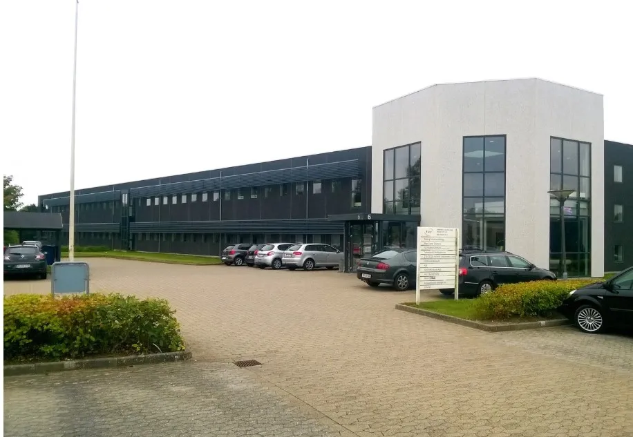 I dette billede er der en moderne bygning med mange vinduer og en stor parkeringsplads med fire parkerede biler og et