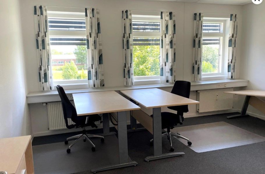 Kontor med to skriveborde og stol, der er placeret under tre vinduer med gardiner. Der er en radiator under et af vinduerne.