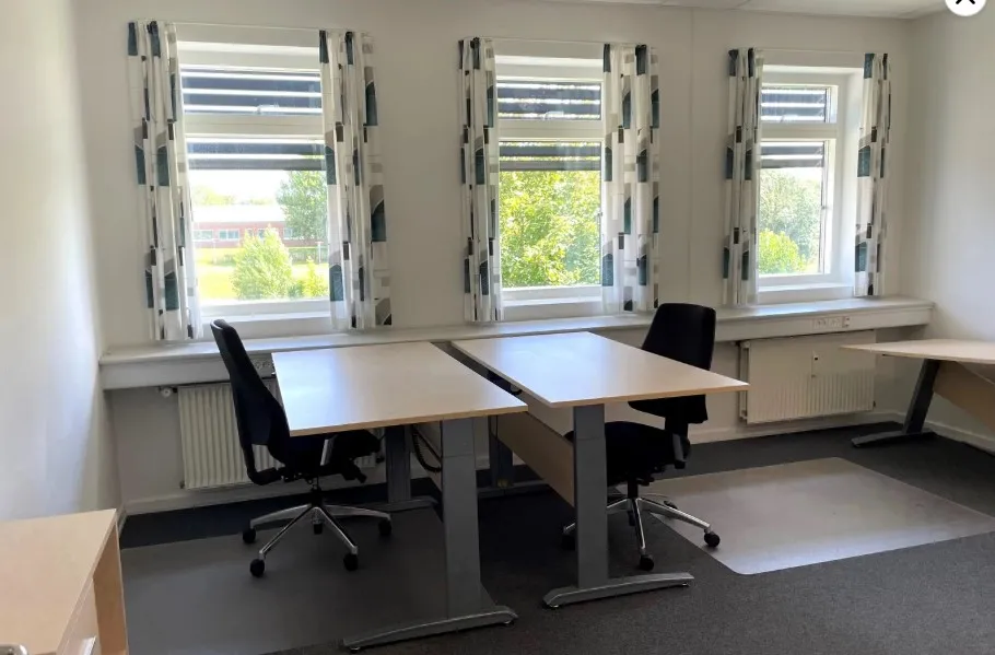 Kontor med to skriveborde og stol, der er placeret under tre vinduer med gardiner. Der er en radiator under et af vinduerne.