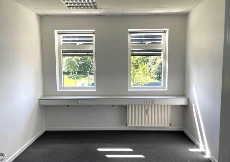 To vinduer i et kontorlokal med hvidt loft, vægge og gulv. To lyse stråler fra solen trænger ind gennem vinduet. Under