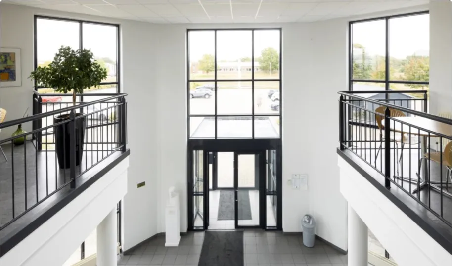 Indgangsparti til kontorlokaler i bygningen med dobbelt glasdør og balkoner med træborde og stole samt en plante
