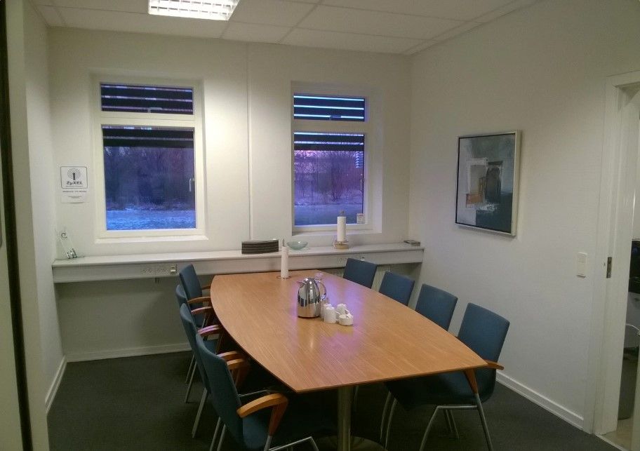 Et kontormødelokale med møbler og udsigt til skoven udenfor vinduer med solblender. En kop og en tekande er placeret på