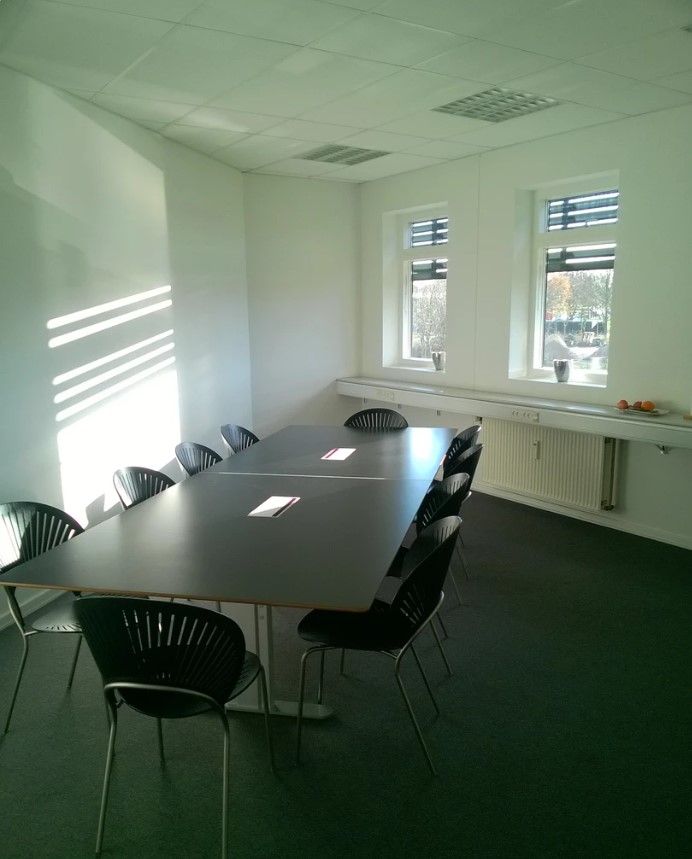 Kontor med to lange borde, flere møbler og to vinduer med gardiner, der lar solen skinne ind i lokalet