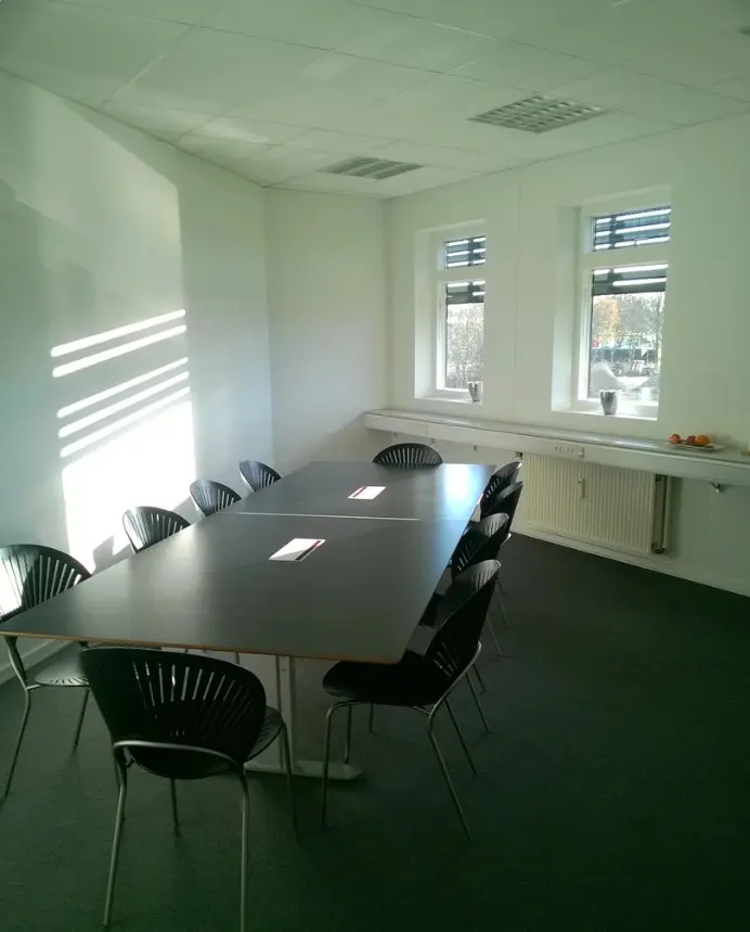 Kontor med to lange borde, flere møbler og to vinduer med gardiner, der lar solen skinne ind i lokalet