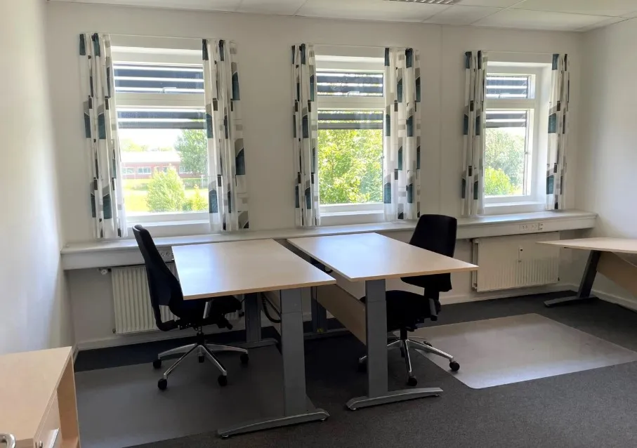To skriveborde og to kontorstolar er placeret i et kontorlokale med tre store vinduer og lysegardiner.