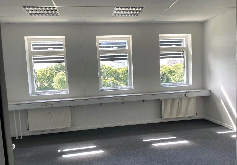 Et tomt kontor med tre vinduer og hvide vægge, gulv og loft. Der er tre radiatorer under vinduet og flere stikkontakter.