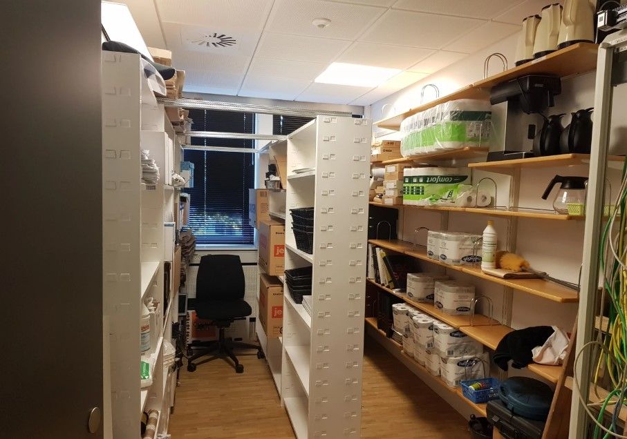 Kontorlokale med høje hylder, stol, kaffemaskine, papkasser, papir, plastposer og tomme bøsser.
