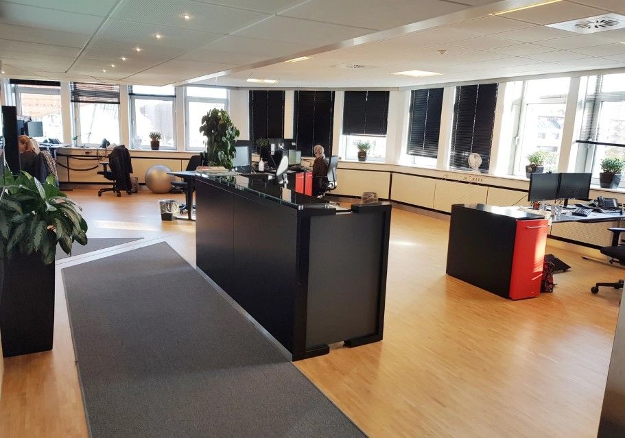 Kontorlokale med trægulv og lyse vinduer, flere skriveborde og stole, plantepot med grønne planter og computere på bordene