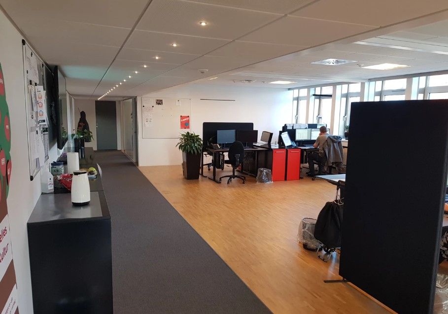 Kontorlokale med trægulv, bord med computer, vase og flere stole. Der er en person siddende og arbejder i et kontorlokale