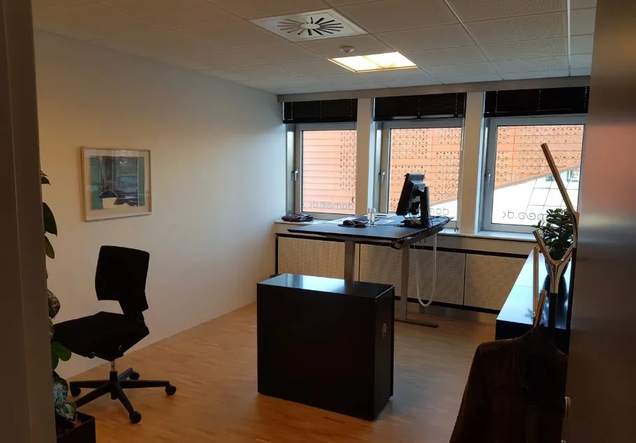 Kontorlokale med skrivebord, stol, plante og computer på skrivebordet, samt vinduer med solskærme og en åben dør til