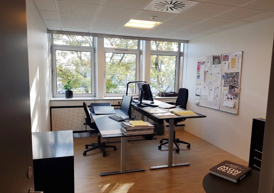 Kontorlokale med to skriveborde, hver med computer og søjle. Plante på vindueskarm og aviser på væggen. Møbler og udstyr til