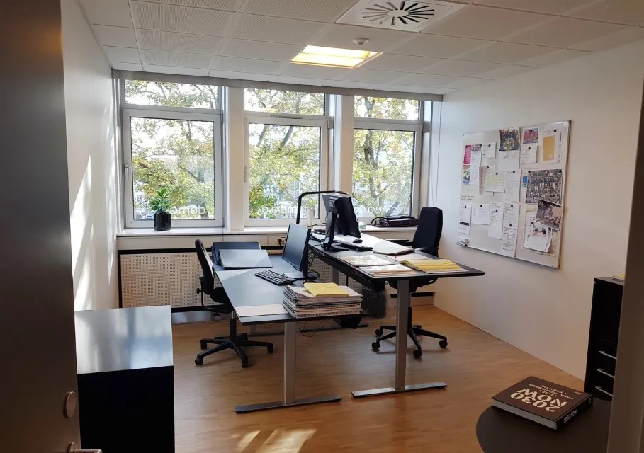 Kontorlokale med to skriveborde, hver med computer og søjle. Plante på vindueskarm og aviser på væggen. Møbler og udstyr til