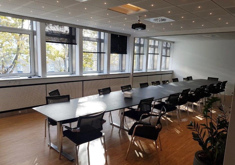 Rummelig kontor med trægulv og store vinduer. To lange borde med sorte stole samt plante i hjørnet. Projektor hængende fra
