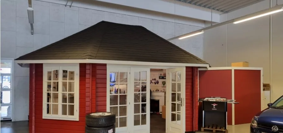 En rødt og hvidt bygget værksted med højt loft og glasdøre og vinduer, der giver god synlighed til bilværkstedet og