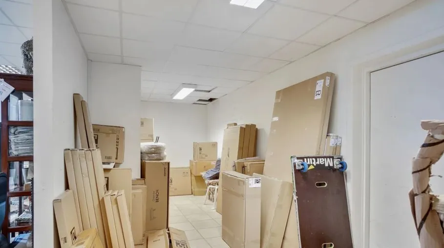 Lager med flere kartoner og trækasser. Store kartoner står opstillet mod væggen. Lyserød og blå skilt er skråt på en kasse.