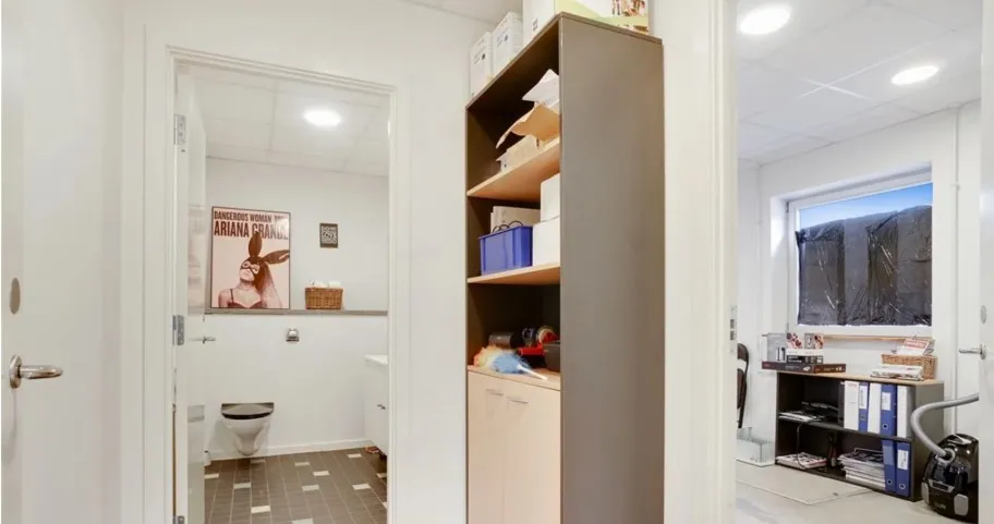 En stor indgangsdør med håndtag og et toilet i en hvid og brun farvetone. Højre side har et højt skab og et skrivebord med