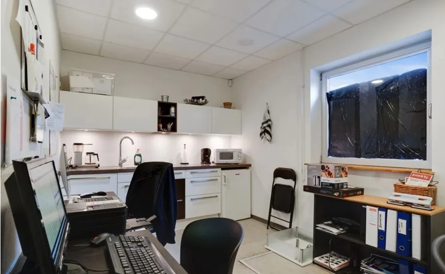 Kontor med højloft, monitor, skrivebord, computer, to stol, mikrobølgeovn, og mange sager på hylder og et vindue