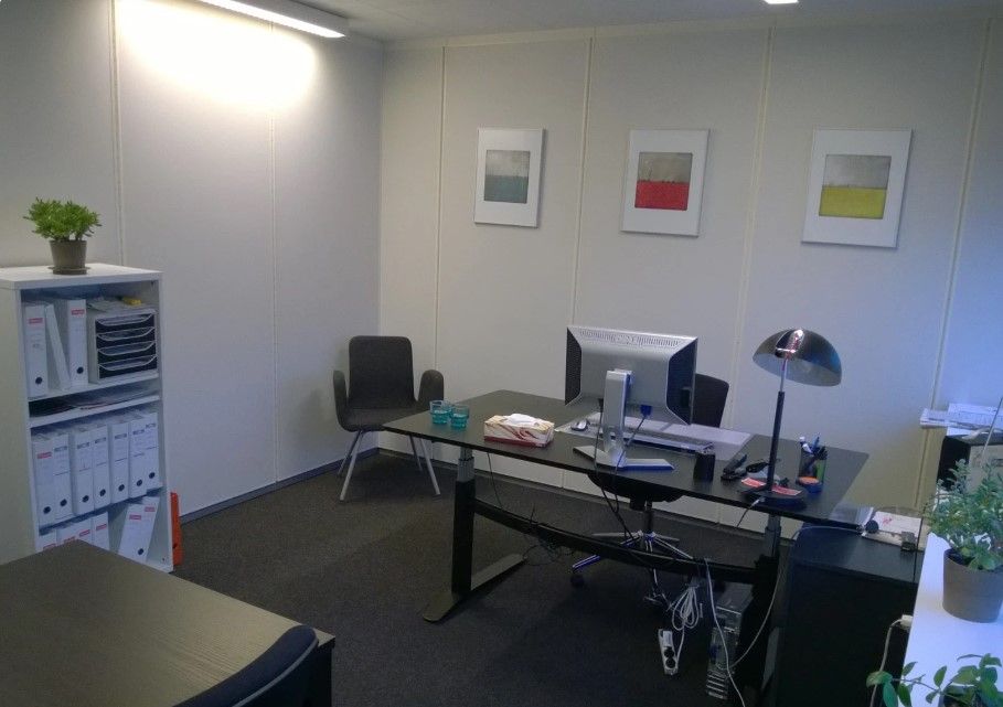 Kontor med højt gulv, højt loft, en bord, to stole, computer, lampe, tre billeder og to potter med grønne planter.