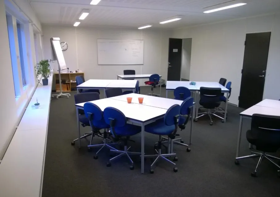 Kontorlokaler med lyse loftslampe, skriveborde, lyseblå stole og et whiteboard bagved