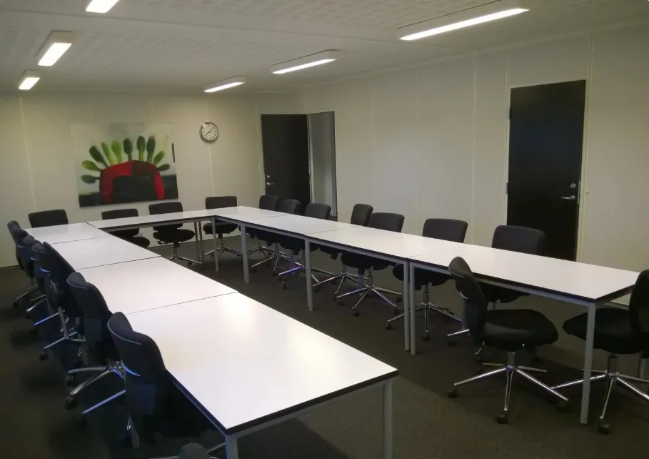 Kontorlokaler med 2 lange borde og flere stole. Rundt om bordene er der små høje stole. På væggen er der et maleri og en