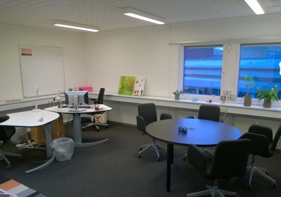 Kontor med to hvide borde, hver med computer og papirkurv, og fire mødestolhede omkring et rundt bord.