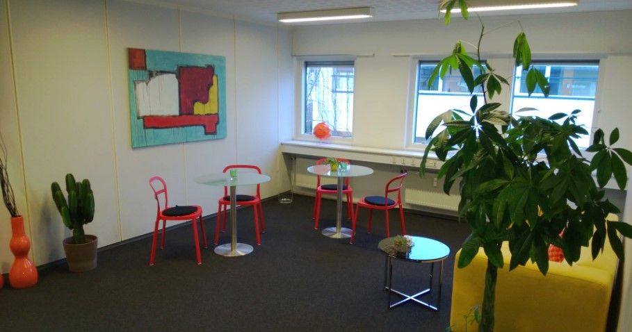 Kontorlokaler med fire borde og stole, lysegul væg, hvide loftslampe, gulv og fire vinduer. Planter og maleri på væggen.