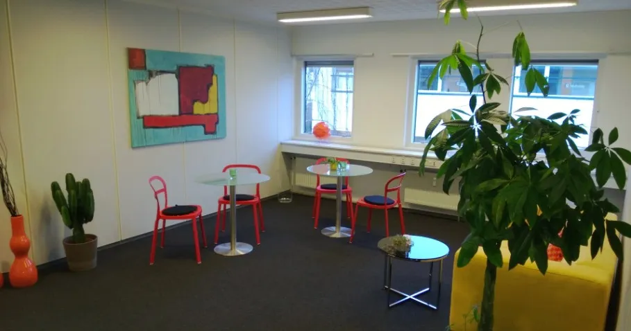 Kontorlokaler med fire borde og stole, lysegul væg, hvide loftslampe, gulv og fire vinduer. Planter og maleri på væggen.