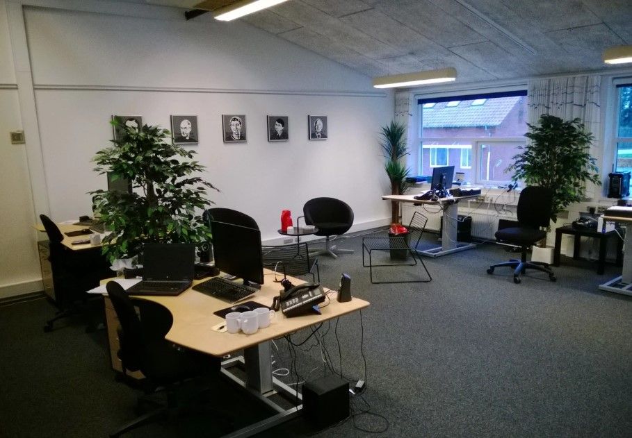 Kontor med fire skriveborde og fire stole samt en plante og en radiator i hjørnet