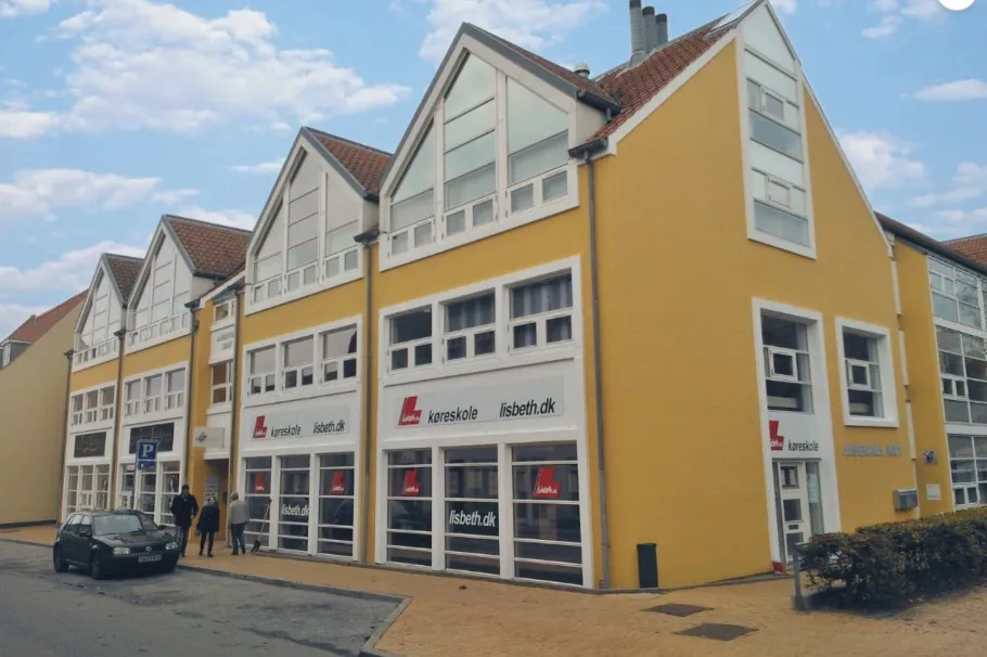 En gul bygning med flere vinduer og tekst i hvide rammer, med folk, der går ind og ud af dørene