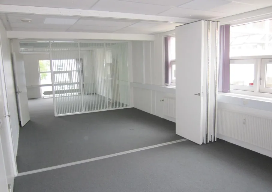 En stor, tom kontoravdeling med hvide vægge, højt til loftet, store vinduer og lyse loftspaneler.