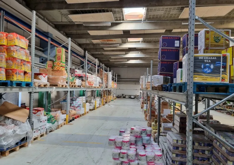 Rummeligt lager med gulv- og højreoler med diverse varer, tøj, emballage og plastposer i forgrunden og baggrunden