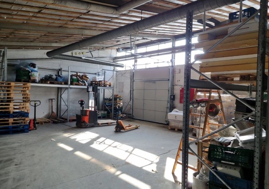 Industriområde med lager og diverse udstyr, træpæle, kørevogne og højrekantet garagedør i lysegråt.