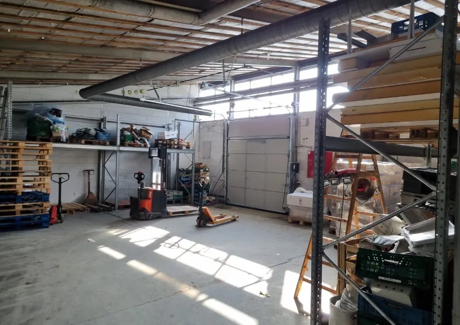 Industriområde med lager og diverse udstyr, træpæle, kørevogne og højrekantet garagedør i lysegråt.