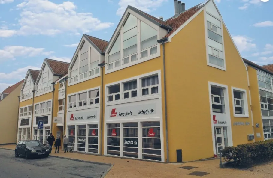 Lejlighed til leje i lysegult bygningskompleks med store vinduer og huse med røde tag. Parkeret bil foran indgangen og folk