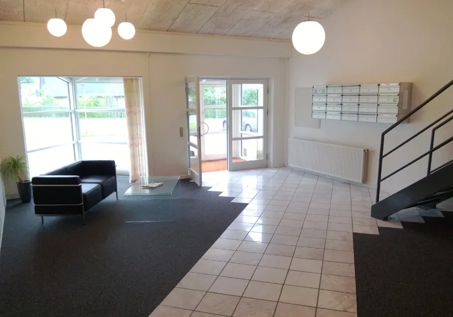 Hvid og lyse interiør med sofa og bord. Styretræppe med skriftpanel og radiator. Glasdøre fører ud til parkeringsplads.