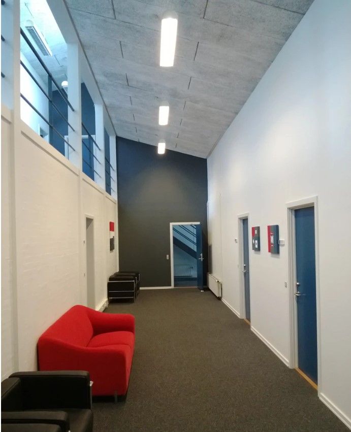 En tom korridor med en høj loftshøjde, lyse gulvbelægning og tre døre, der fører til forskellige rum. En sofa og en stol