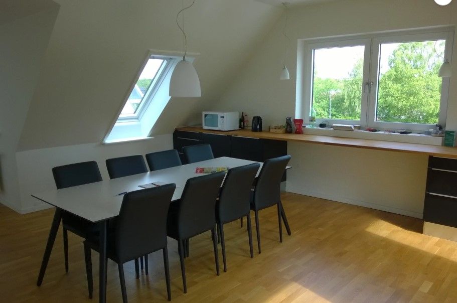 Et lyseværelse med et højt loft, et rektangulært spisebord og stole samt en mikrobølgeovn og vinduer med udsigt til skoven