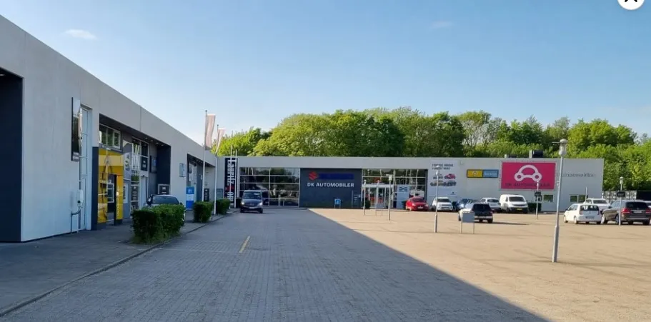Bilforretning med biler og vogne parkeret foran og på indre parkeringsplads. På facaden er der reklamer og tekst som DK