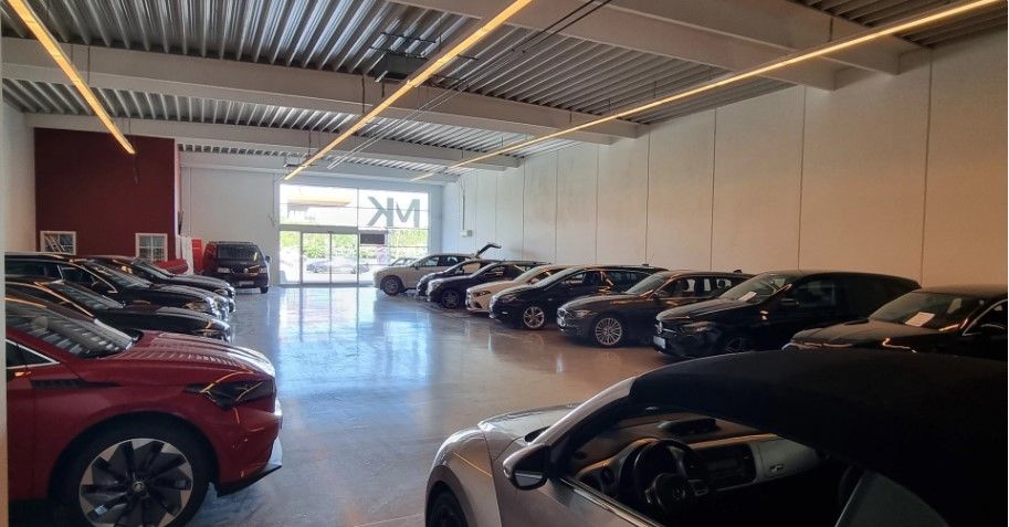 Inde i et showroom er flere biler parkeret, inklusive en rød, en hvid og en sort cabriolet og en gulvmonteret lysekrone