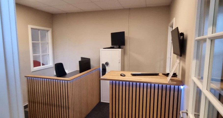 Kontorrum med to skriveborde og stole samt en hvid kabinet med et skærm og en monitor monteret på væggen.