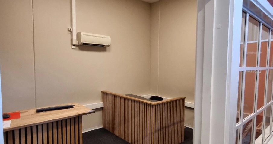 To lysebrune træpulte og en hvid klimaanlægsventilator. Der er også en glasdør med hvide sider og en hvid dørhåndtag. Gulvet