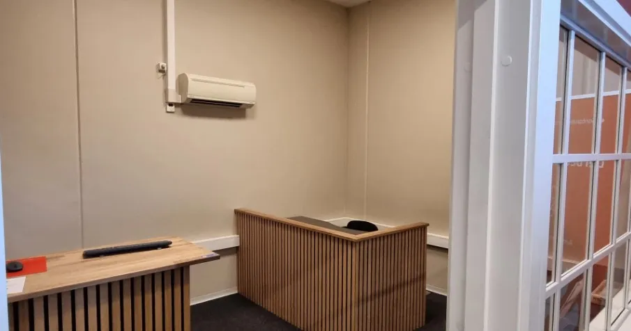 To lysebrune træpulte og en hvid klimaanlægsventilator. Der er også en glasdør med hvide sider og en hvid dørhåndtag. Gulvet