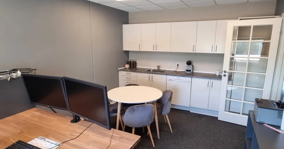 Kontor med høje hvide vægge og lysegulve. Rundt bord med flere stole samt to skærme og tastatur. Hvide skabe med