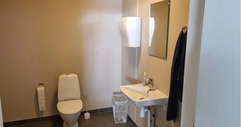 Hjørnebadeværelse med hvid toilet, hængende håndklædeholder og hvidt vask og håndklæde. Højloftet rum med hvidt loft og væg,