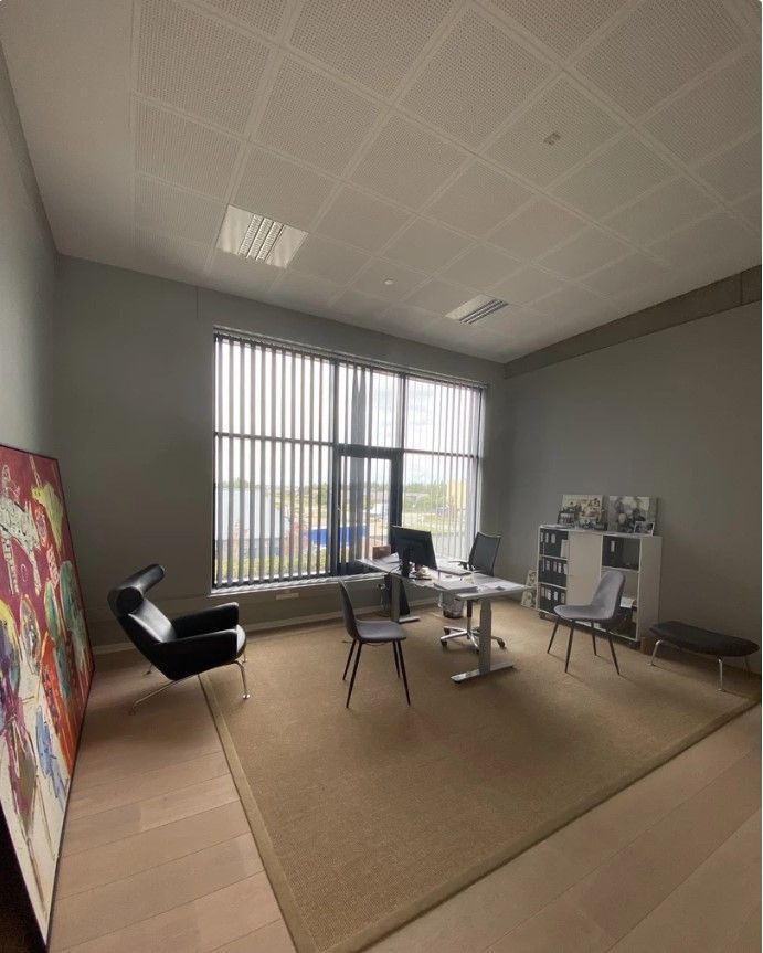 Et tomt kontor med skrivebord, stol, gulvtæppe, maleri og vinduer med persienner